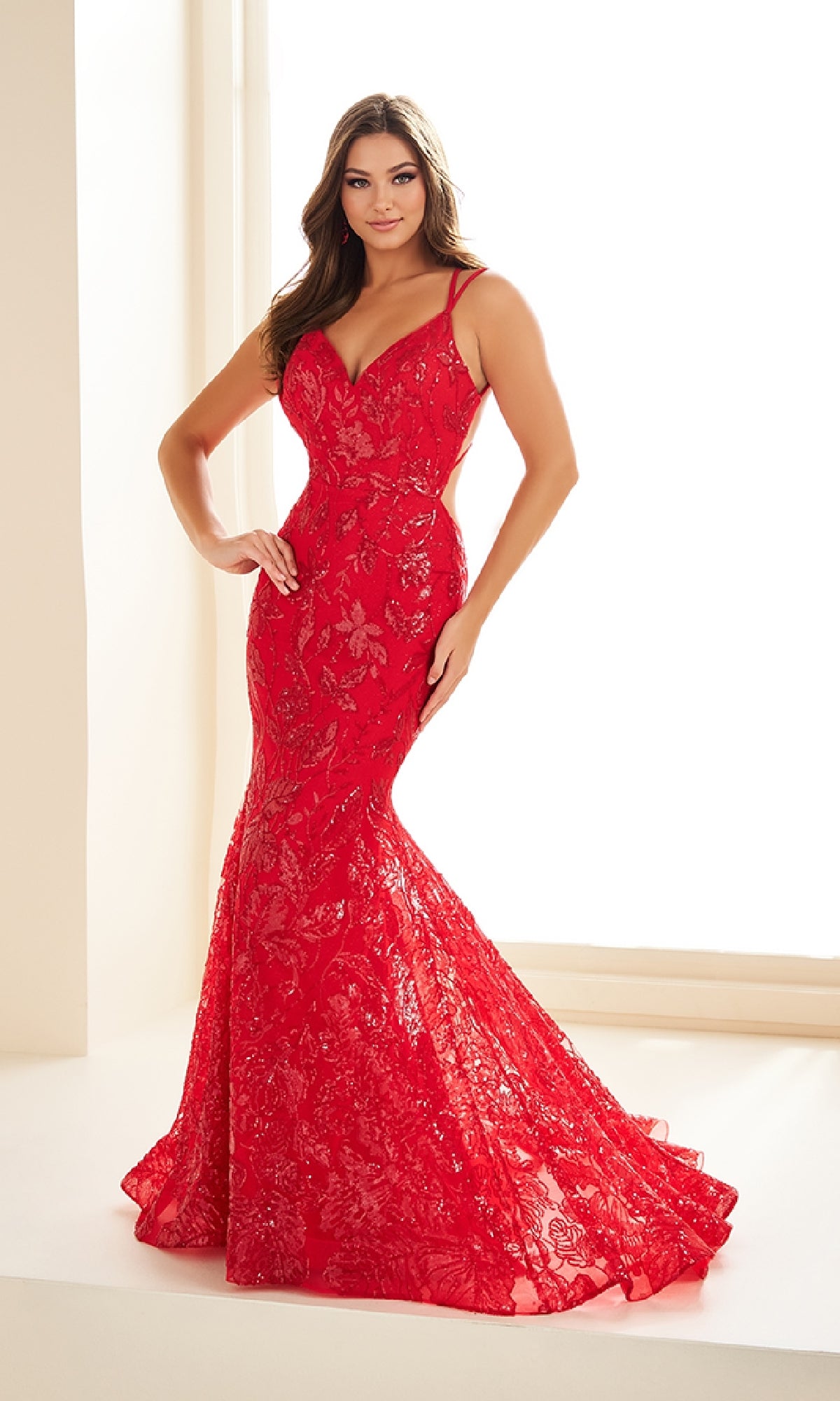 A woman poses elegantly in a well-lit room wearing the Ellie Wilde EW36034, a fitted long prom dress featuring an open back, red lace, thin straps, and a flared hem.